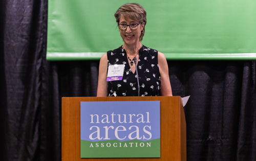Woman standing at a podium speaking to audience at a conference.