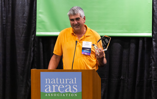 Ken McCarty standing at a podium holding an award, speaking to an audience at a conference.
